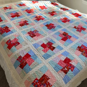 K&ouml;nnte beinhalten: Eine Patchwork-Decke mit wei&szlig;em Hintergrund und Quadraten aus rotem, rosa, blauem und gr&uuml;nem Stoff. Die Decke ist aus verschiedenen Mustern und Texturen gefertigt.