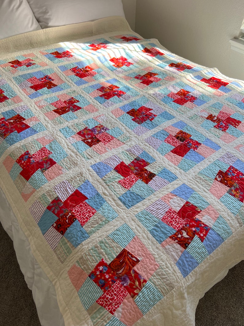 K&ouml;nnte beinhalten: Eine Patchwork-Decke mit wei&szlig;em Hintergrund und Quadraten aus rotem, rosa, blauem und gr&uuml;nem Stoff. Die Decke ist aus verschiedenen Mustern und Texturen gefertigt.