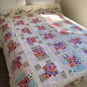 以下が含まれることがあります： ピンク、パープル、オレンジ、ブルーの色の花柄が施された白いキルト。キルトは正方形で構成されており、ダイヤモンドパターンでキルティングされています。