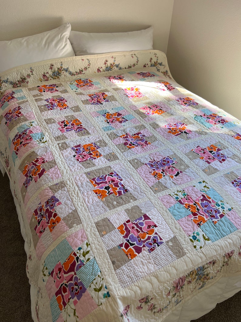 以下が含まれることがあります： ピンク、パープル、オレンジ、ブルーの色の花柄が施された白いキルト。キルトは正方形で構成されており、ダイヤモンドパターンでキルティングされています。