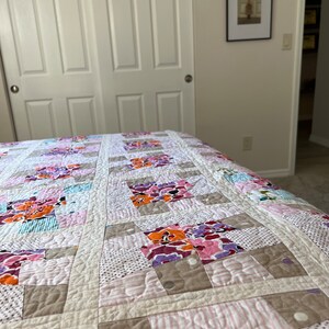 以下が含まれることがあります： ピンク、オレンジ、パープル、ブルーの色合いのさまざまな花柄の入ったパッチワークキルト。キルトは白い背景で作られており、キルティングされた質感があります。
