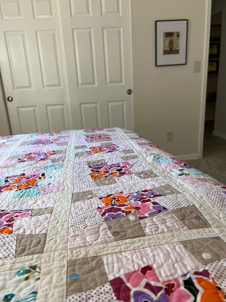 以下が含まれることがあります： ピンク、オレンジ、パープル、ブルーの色合いのさまざまな花柄の入ったパッチワークキルト。キルトは白い背景で作られており、キルティングされた質感があります。