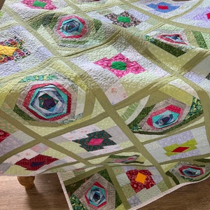 Peut inclure: Une courtepointe en patchwork vert et blanc avec un motif floral. La courtepointe est compos&eacute;e de carr&eacute;s et de rectangles de tissus diff&eacute;rents, et les fleurs sont faites de carr&eacute;s et de rectangles plus petits. La courtepointe est pli&eacute;e en deux et repose sur un sol en bois.