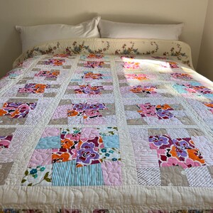 以下が含まれることがあります： ピンク、パープル、オレンジ、ブルーの色合いの花柄のキルト。キルトはコットンとリネンなど、さまざまな生地で作られています。キルトは白い枕カバーのあるベッドの上に置かれています。