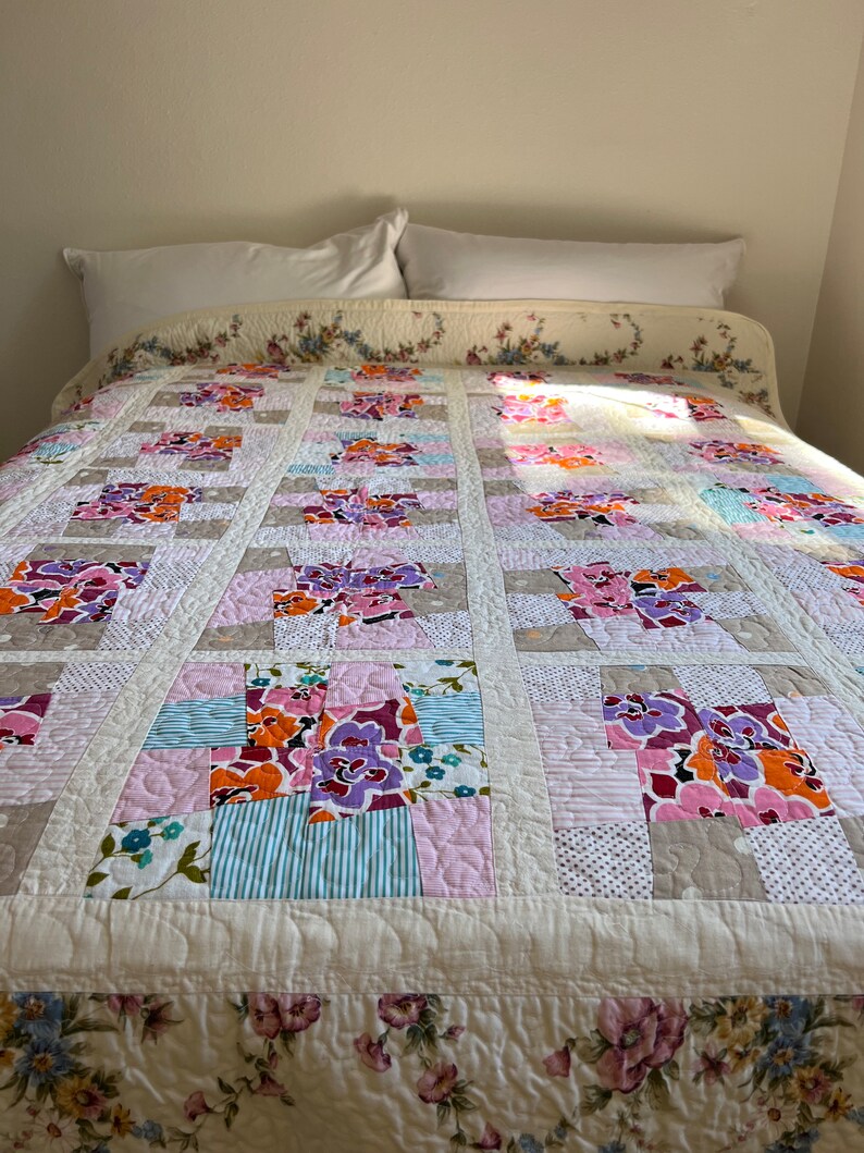 以下が含まれることがあります： ピンク、パープル、オレンジ、ブルーの色合いの花柄のキルト。キルトはコットンとリネンなど、さまざまな生地で作られています。キルトは白い枕カバーのあるベッドの上に置かれています。