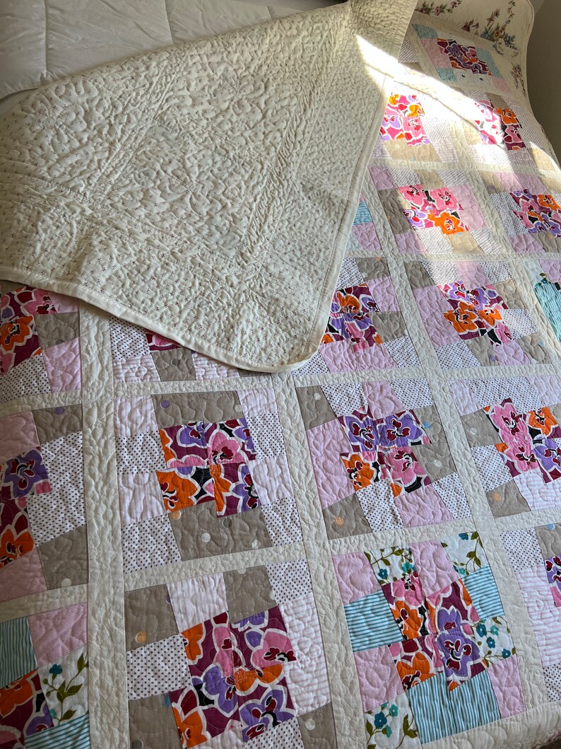 以下が含まれることがあります： 花柄の白いキルト。キルトは異なる生地の正方形で構成されており、それぞれ異なる花柄のデザインが施されています。キルトは半分に折り畳まれ、上半分は下半分に折り畳まれています。