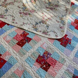 K&ouml;nnte beinhalten: Eine wei&szlig;e Bettdecke mit einem Blumenmuster auf der einen Seite und einem Patchworkmuster auf der anderen Seite. Die Patchworkseite besteht aus Quadraten aus rotem, rosa, blauem und gr&uuml;nem Stoff.