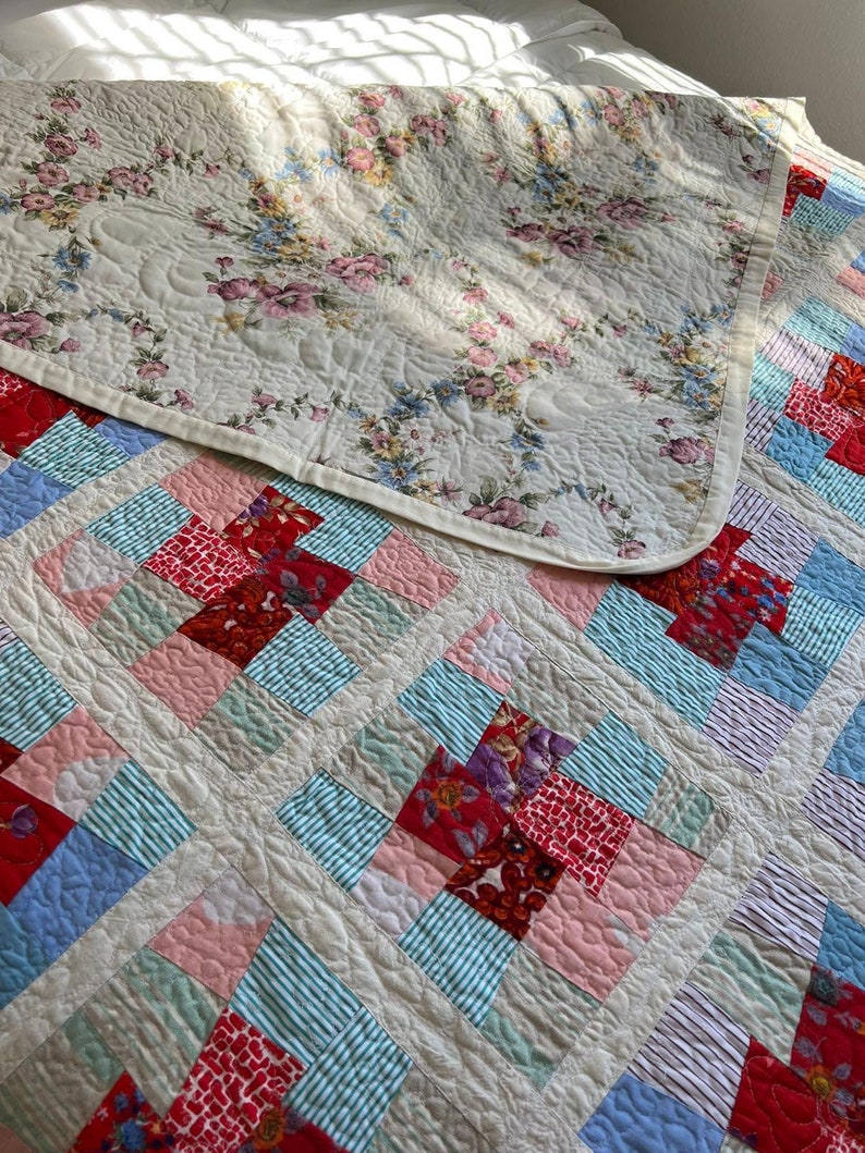 K&ouml;nnte beinhalten: Eine wei&szlig;e Bettdecke mit einem Blumenmuster auf der einen Seite und einem Patchworkmuster auf der anderen Seite. Die Patchworkseite besteht aus Quadraten aus rotem, rosa, blauem und gr&uuml;nem Stoff.
