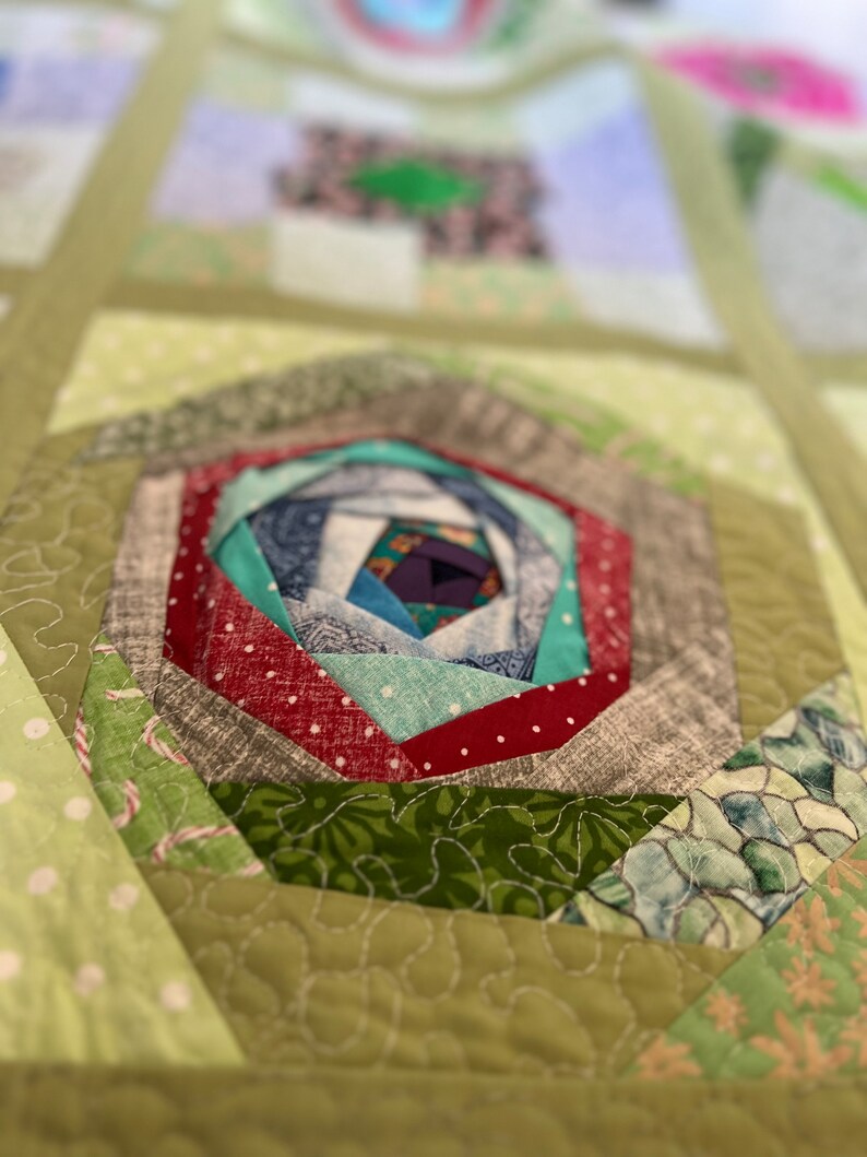 Peut inclure: Gros plan sur une courtepointe verte et blanche avec un motif en spirale au centre. La spirale est faite de diff&eacute;rents tissus, dont du rouge, du vert, du bleu et du blanc. La courtepointe est matelass&eacute;e avec un fil blanc.