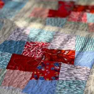 K&ouml;nnte beinhalten: Eine Patchwork-Decke mit verschiedenen roten, blauen und wei&szlig;en Stoffen. Die Decke besteht aus Quadraten und Rechtecken, die N&auml;hte sind sichtbar. Die Decke ist mit einem einfachen Stich gesteppt.