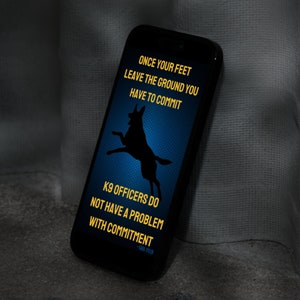May include: A black smartphone with a blue background and a silhouette of a German Shepherd dog jumping. The text on the screen reads "Once your feet leave the ground you have to commit. K9 officers do not have a problem with commitment. -Sara Evler"