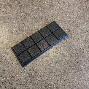 May include: A black rectangular object with a textured surface, divided into twelve square sections. It has a raised border and is placed on a speckled grey surface. The squares have a rough, granular texture.