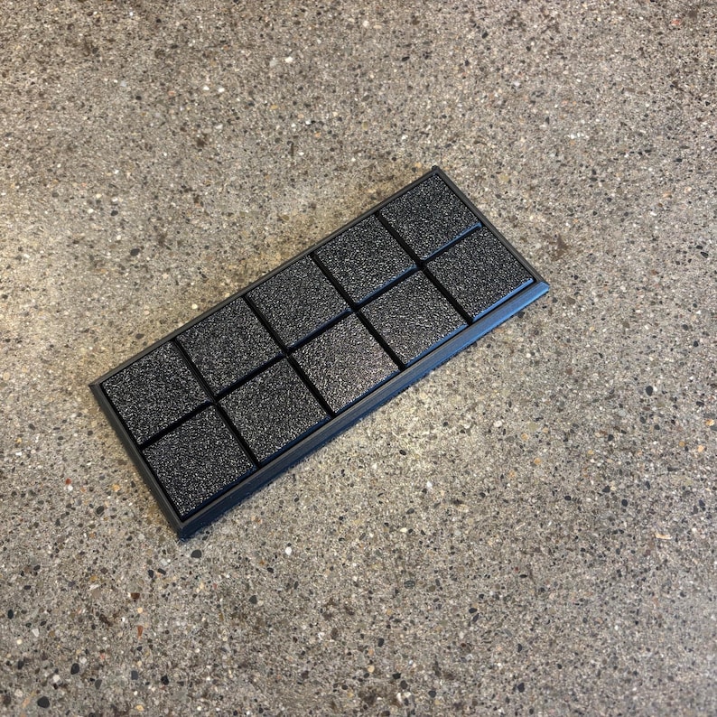May include: A black rectangular object with a textured surface, divided into twelve square sections. It has a raised border and is placed on a speckled grey surface. The squares have a rough, granular texture.