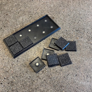 May include: A black rectangular tray with embedded magnets and several square tiles with speckled surfaces. Some tiles are within the tray, while others are scattered nearby. The tray and tiles appear to be made of plastic.