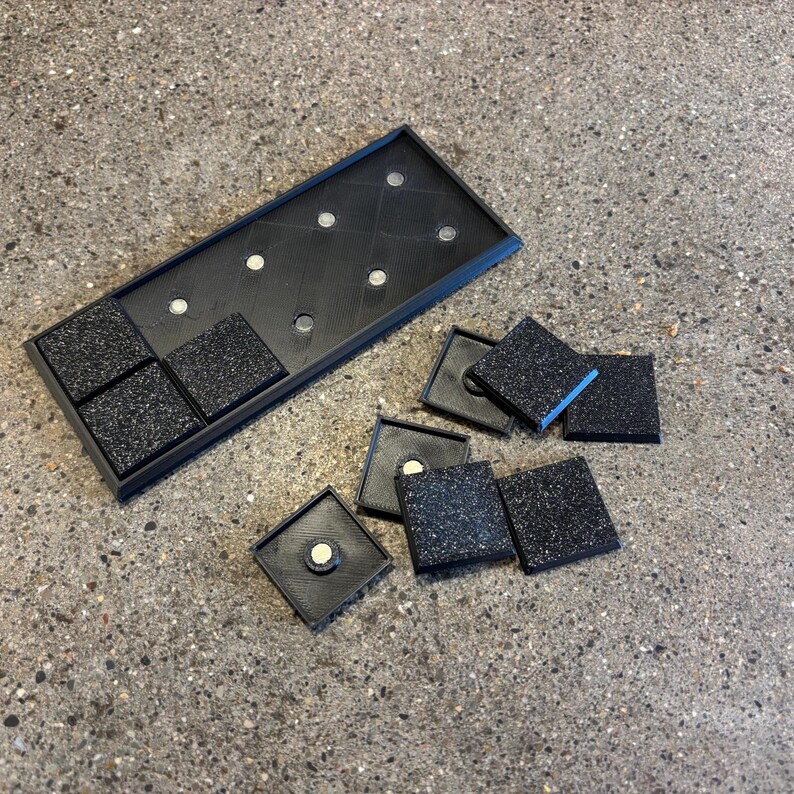 May include: A black rectangular tray with embedded magnets and several square tiles with speckled surfaces. Some tiles are within the tray, while others are scattered nearby. The tray and tiles appear to be made of plastic.
