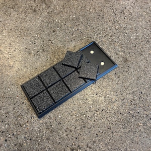 May include: A black tray with nine square compartments, each containing a textured, black square. Several additional squares are placed on top of the tray. The tray is open, revealing a black interior with two white dots.