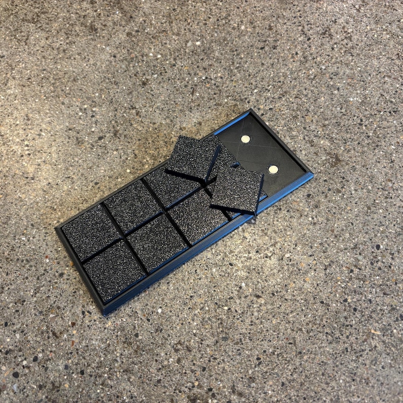 May include: A black tray with nine square compartments, each containing a textured, black square. Several additional squares are placed on top of the tray. The tray is open, revealing a black interior with two white dots.