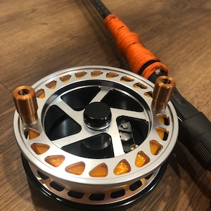 May include: A silver fly fishing reel with an orange line and black accents. The reel is attached to a black and orange fly fishing rod.