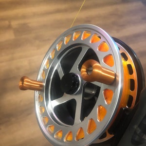 Può includere: Un mulinello da pesca a mosca argentato con una bobina arancione e una maniglia color rame. Il mulinello è avvolto con filo da pesca a mosca arancione.