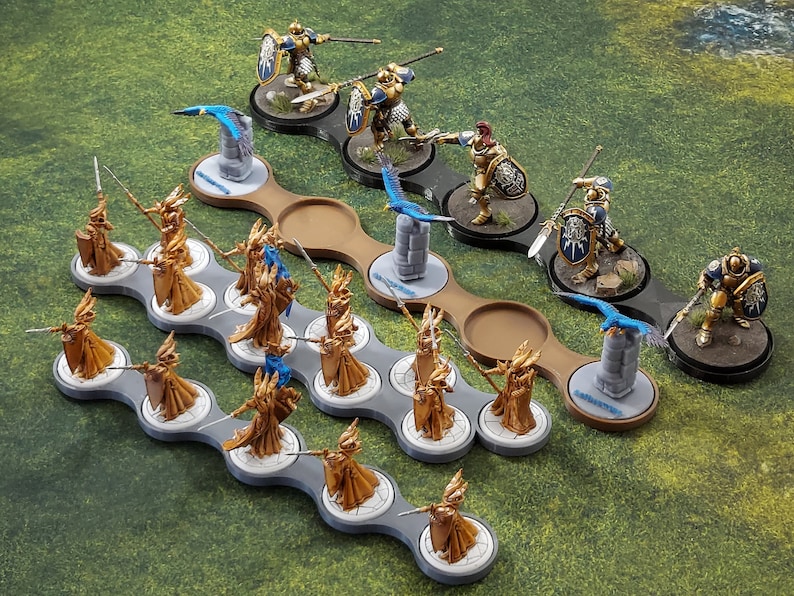 May include: A tabletop wargaming set featuring miniature figures of warriors clad in gold armour. The figures are arranged on circular bases and are positioned on a grey and brown plastic tray. The figures are wielding swords and spears. There are also blue and white miniature birds on the tray.