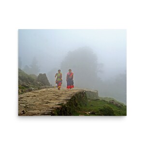 Puede incluir: Dos personas caminan por un sendero de piedra en un paisaje montañoso brumoso. La persona que va delante lleva un vestido estampado de colores y un bastón. La persona que va detrás lleva un sari rojo y azul.