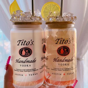 K&ouml;nnte beinhalten: Zwei durchsichtige Glasbeh&auml;lter mit Deckeln, gef&uuml;llt mit Eis und Zitronenscheiben. Die Beh&auml;lter tragen ein Etikett mit der Aufschrift "Tito's Handmade Vodka" und darunter die Worte "Austin, Texas".