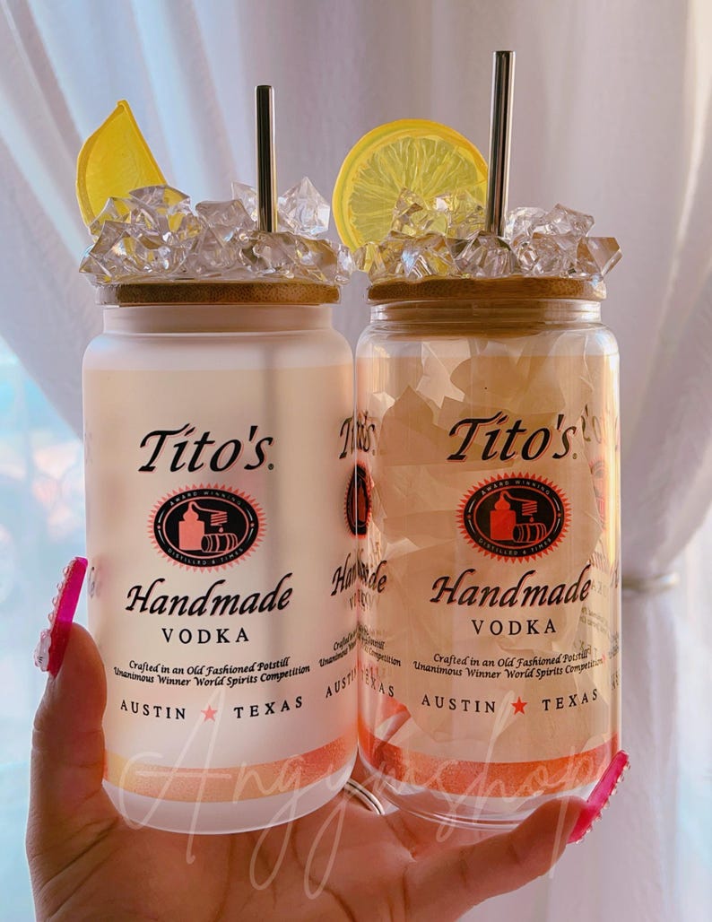 K&ouml;nnte beinhalten: Zwei durchsichtige Glasbeh&auml;lter mit Deckeln, gef&uuml;llt mit Eis und Zitronenscheiben. Die Beh&auml;lter tragen ein Etikett mit der Aufschrift "Tito's Handmade Vodka" und darunter die Worte "Austin, Texas".