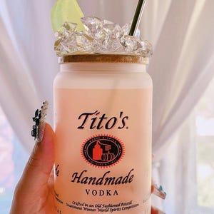 K&ouml;nnte beinhalten: Eine gefrostete Glasflasche Tito's Handmade Vodka mit einem Limettenst&uuml;ck und Eis. Die Flasche hat ein rotes Etikett mit dem Text "Tito's Handmade Vodka" und "Crafted in an Old Fashioned Potstill Unanimous Winner World Spirits Competition Austin Texas".