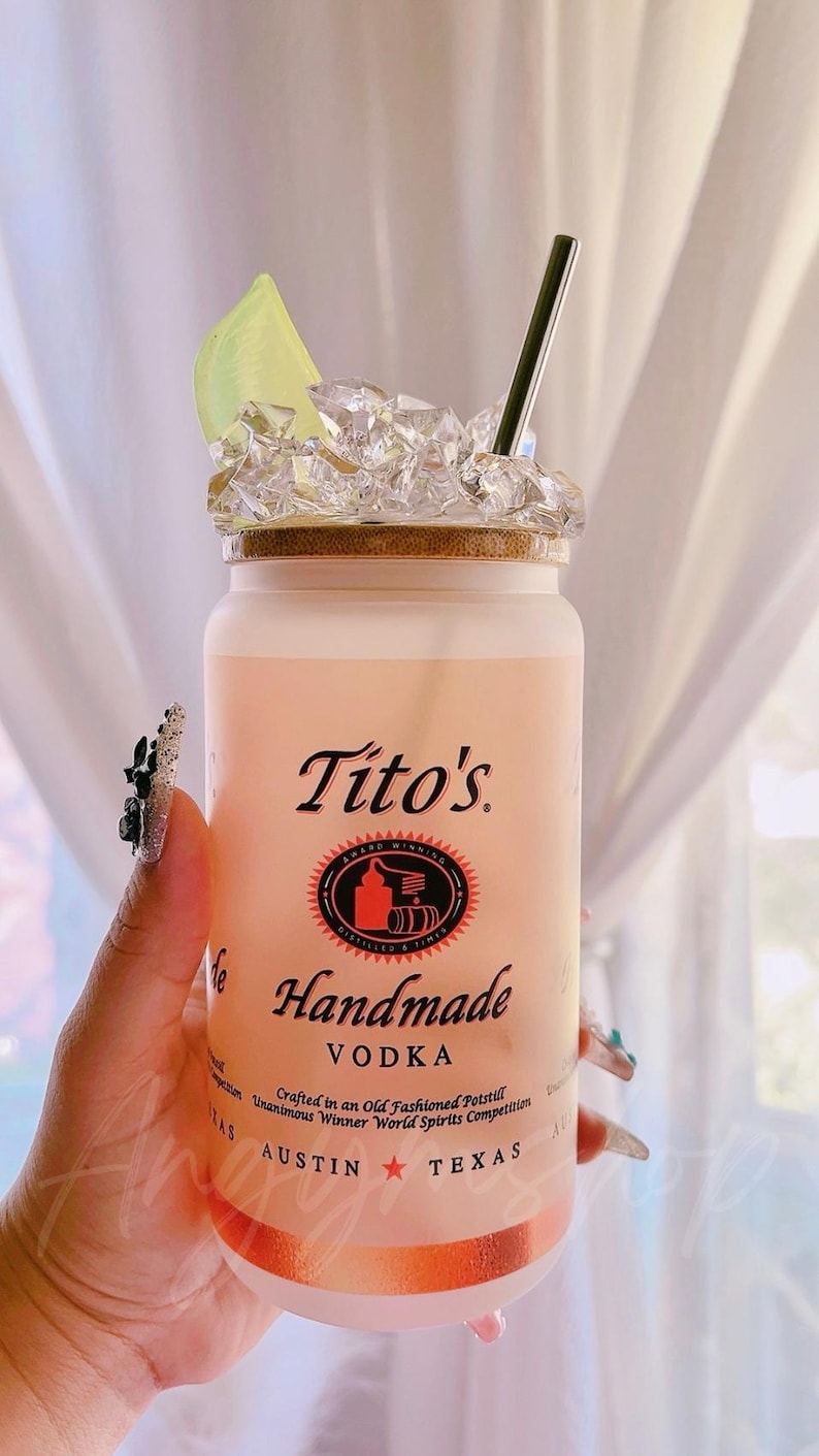 K&ouml;nnte beinhalten: Eine gefrostete Glasflasche Tito's Handmade Vodka mit einem Limettenst&uuml;ck und Eis. Die Flasche hat ein rotes Etikett mit dem Text "Tito's Handmade Vodka" und "Crafted in an Old Fashioned Potstill Unanimous Winner World Spirits Competition Austin Texas".