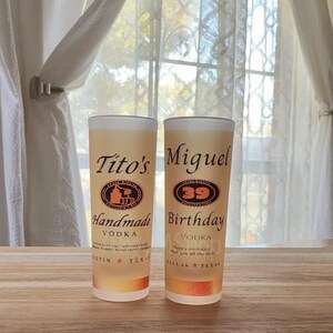 May include: Two tall, frosted glasses with a light amber liquid inside. One glass reads "Tito's Handmade Vodka, Austin Texas." The other says "Miguel 39 Birthday, Dallas Texas." Both glasses have a white rim.