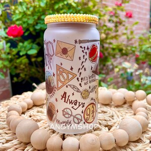 Op de afbeelding: Een witte herbruikbare waterfles met een Harry Potter-thema. De fles heeft verschillende Harry Potter-symbolen en -personages, waaronder het symbool van de Doodsgeesten, een gouden snitch en de woorden "Always" en "Muggle". De fles heeft een gouden glitterdeksel.