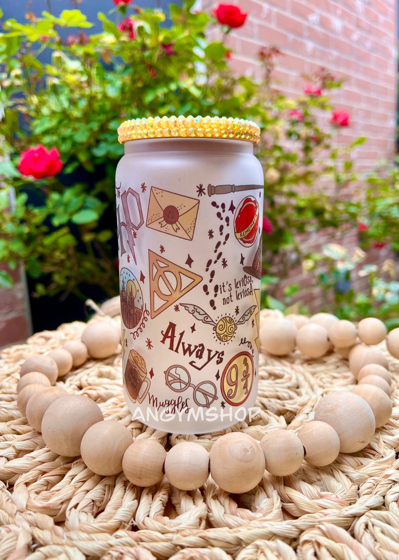 Op de afbeelding: Een witte herbruikbare waterfles met een Harry Potter-thema. De fles heeft verschillende Harry Potter-symbolen en -personages, waaronder het symbool van de Doodsgeesten, een gouden snitch en de woorden "Always" en "Muggle". De fles heeft een gouden glitterdeksel.