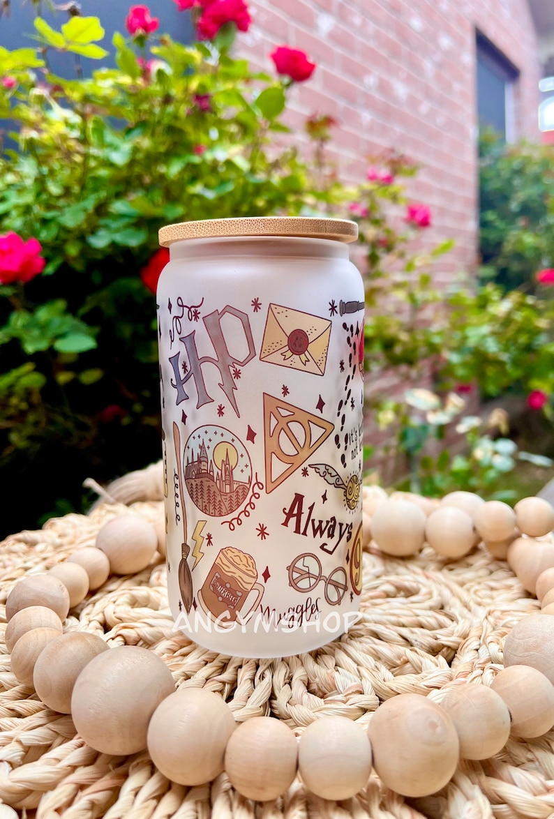 Op de afbeelding: Een witte, gematteerde glazen pot met een bamboe deksel met een Harry Potter-thema. De pot is versierd met illustraties van het kasteel van Zweinstein, het symbool van de Doodsgeesten, een bliksem, een gouden snitch, een rond brilletje, een Zweinstein-brief en de woorden "Always".