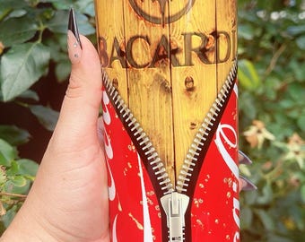 May include: A stainless steel tumbler with a wooden-look design featuring the Bacardi logo. The lower half of the tumbler has a red zipper design with white accents. The tumbler is held by a person.