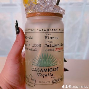 Puede incluir: Una botella de vidrio esmerilado de Casamigos Tequila Blanco, con una rodaja de lima y hielo en la parte superior. La etiqueta de la botella dice "Casamigos Tequila Blanco, Hecho en Jalisco, M&eacute;xico, Numero 23044, Exclusivo".