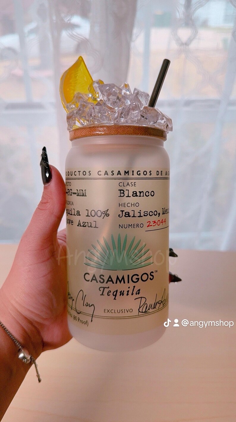 Puede incluir: Una botella de vidrio esmerilado de Casamigos Tequila Blanco, con una rodaja de lima y hielo en la parte superior. La etiqueta de la botella dice "Casamigos Tequila Blanco, Hecho en Jalisco, M&eacute;xico, Numero 23044, Exclusivo".