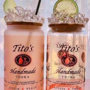 K&ouml;nnte beinhalten: Zwei durchsichtige Glasbeh&auml;lter mit braunem Deckel und wei&szlig;em Etikett. Das Etikett tr&auml;gt den Schriftzug "Tito's Handmade Vodka" mit einem roten Logo und dem Text "Crafted in an Old Fashioned Potstill Unanimous Winner World Spirits Competition Austin Texas".