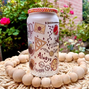 Op de afbeelding: Een witte glazen beker met een rood, oranje en geel ontwerp met illustraties met Harry Potter-thema, waaronder het Hogwarts-wapen, een gouden snitch en de woorden "Always" en "Muggle".