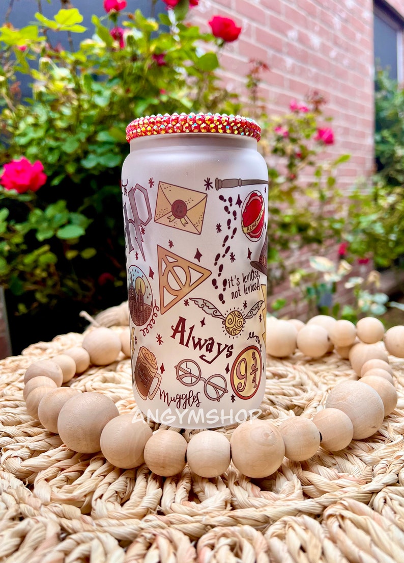Op de afbeelding: Een witte glazen beker met een rood, oranje en geel ontwerp met illustraties met Harry Potter-thema, waaronder het Hogwarts-wapen, een gouden snitch en de woorden "Always" en "Muggle".