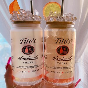 K&ouml;nnte beinhalten: Zwei klare Glasbecher mit mattem Finish, jeweils mit einem Strohhalm und einer Orangenscheibe obenauf. Die Becher sind mit Eis gef&uuml;llt und haben einen braunen Holzdeckel. Die Becher sind mit "Tito's Handmade Vodka" beschriftet, mit dem Text "Crafted in an Old Fashioned Pot Still Unanimous Winner World Spirits Competition Austin Texas".