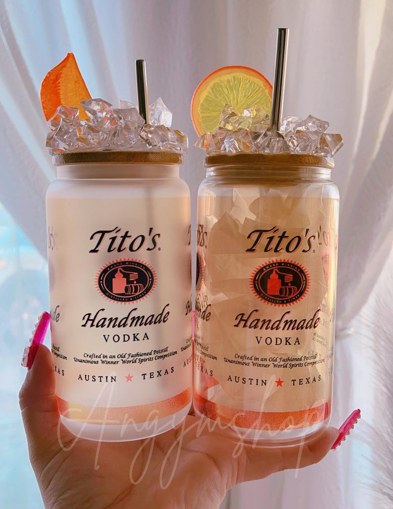 K&ouml;nnte beinhalten: Zwei klare Glasbecher mit mattem Finish, jeweils mit einem Strohhalm und einer Orangenscheibe obenauf. Die Becher sind mit Eis gef&uuml;llt und haben einen braunen Holzdeckel. Die Becher sind mit "Tito's Handmade Vodka" beschriftet, mit dem Text "Crafted in an Old Fashioned Pot Still Unanimous Winner World Spirits Competition Austin Texas".