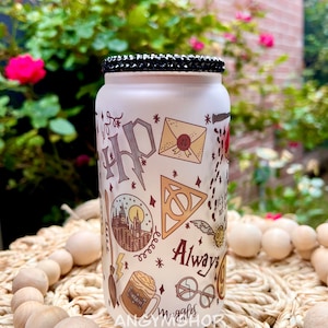 Op de afbeelding: Een witte, gematteerde glazen pot met een zwarte strass-deksel. De pot is versierd met een Harry Potter-thema, waaronder het Hogwarts-wapen, een gouden snitch, een bliksemflits en de woorden "Always".