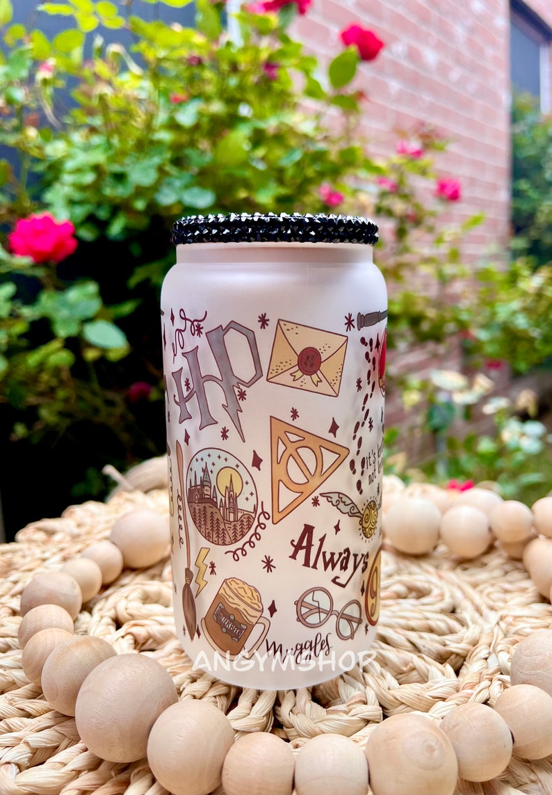 Op de afbeelding: Een witte, gematteerde glazen pot met een zwarte strass-deksel. De pot is versierd met een Harry Potter-thema, waaronder het Hogwarts-wapen, een gouden snitch, een bliksemflits en de woorden "Always".