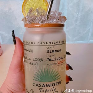 Puede incluir: Una botella de vidrio esmerilado con una tapa de madera y una pajita. La botella est&aacute; llena de hielo y una rodaja de lim&oacute;n. La botella tiene una etiqueta que dice "Casamigos Tequila" y "Blanco" en texto negro. La etiqueta tambi&eacute;n dice "Hecho en Jalisco, M&eacute;xico" y "Numero 23044".