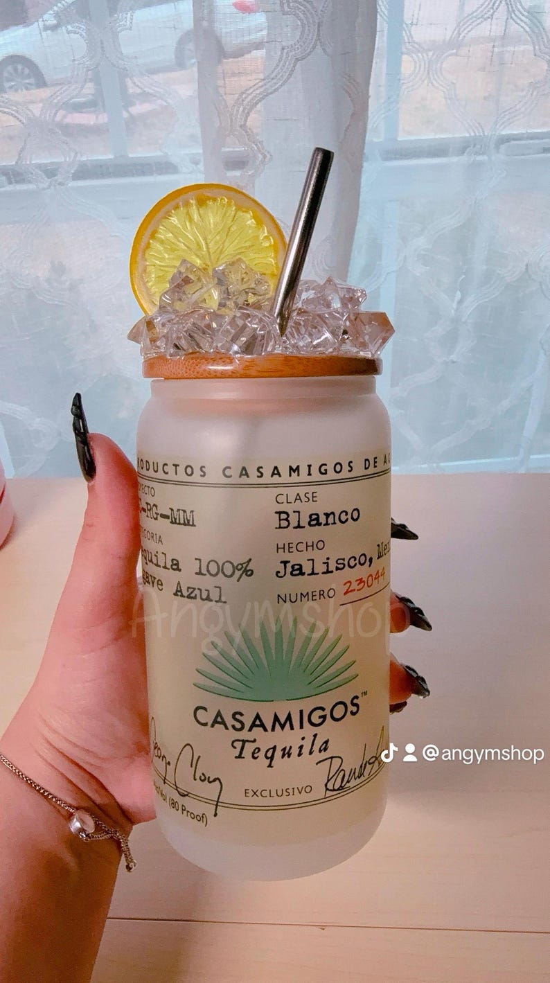 Puede incluir: Una botella de vidrio esmerilado con una tapa de madera y una pajita. La botella est&aacute; llena de hielo y una rodaja de lim&oacute;n. La botella tiene una etiqueta que dice "Casamigos Tequila" y "Blanco" en texto negro. La etiqueta tambi&eacute;n dice "Hecho en Jalisco, M&eacute;xico" y "Numero 23044".