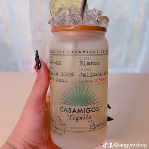 Puede incluir: Una botella de vidrio esmerilado con tapa de bamb&uacute; y una pajita plateada. La botella est&aacute; llena de hielo y una rodaja de lima. La etiqueta de la botella dice "Casamigos Tequila" y "Blanco" en texto verde. La botella est&aacute; sobre una superficie de madera.