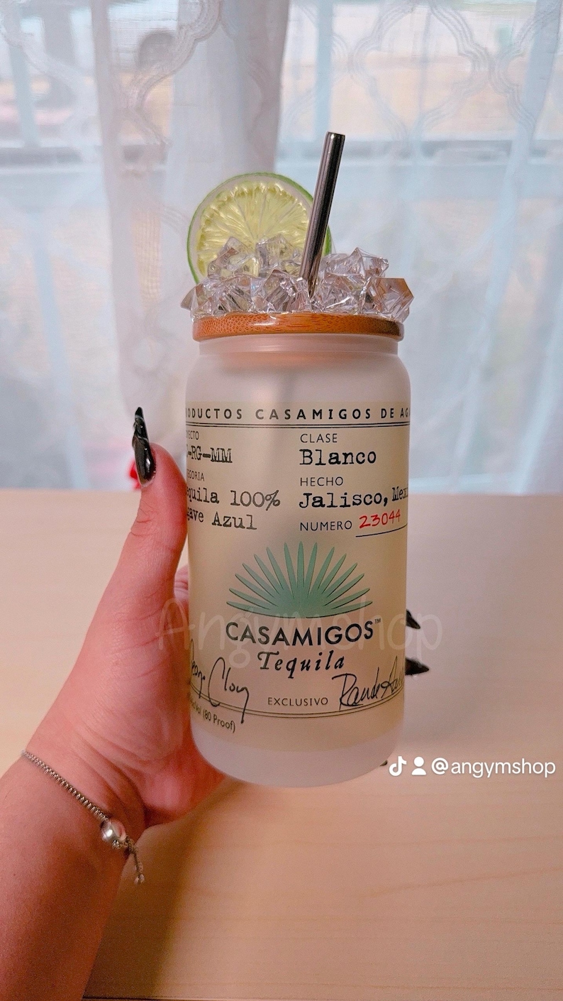 Puede incluir: Una botella de vidrio esmerilado con tapa de bamb&uacute; y una pajita plateada. La botella est&aacute; llena de hielo y una rodaja de lima. La etiqueta de la botella dice "Casamigos Tequila" y "Blanco" en texto verde. La botella est&aacute; sobre una superficie de madera.