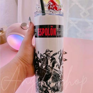 May include: White stainless steel tumbler with a black and white illustration of a skeleton riding a rooster. The tumbler has a black lid and a black straw. The text "Espolón Tequila Blanco" is printed on the tumbler.