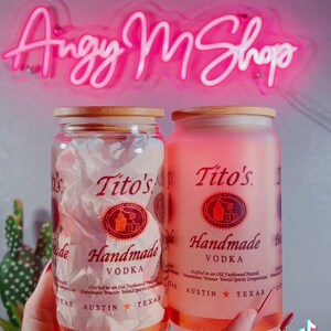 K&ouml;nnte beinhalten: Zwei Glasbeh&auml;lter mit Bambusdeckeln, jeder mit einer klaren Fl&uuml;ssigkeit und einem Etikett mit der Aufschrift "Tito's Handmade Vodka". Die Etiketten sind rot und wei&szlig; mit dem Text "Crafted in an Old Fashioned Pot Still" und "Unanimous Winner World Spirits Competition". Die Beh&auml;lter werden in der Hand einer Person gehalten.