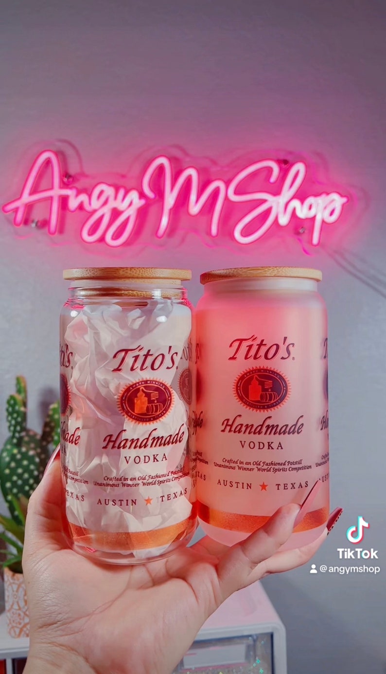 K&ouml;nnte beinhalten: Zwei Glasbeh&auml;lter mit Bambusdeckeln, jeder mit einer klaren Fl&uuml;ssigkeit und einem Etikett mit der Aufschrift "Tito's Handmade Vodka". Die Etiketten sind rot und wei&szlig; mit dem Text "Crafted in an Old Fashioned Pot Still" und "Unanimous Winner World Spirits Competition". Die Beh&auml;lter werden in der Hand einer Person gehalten.